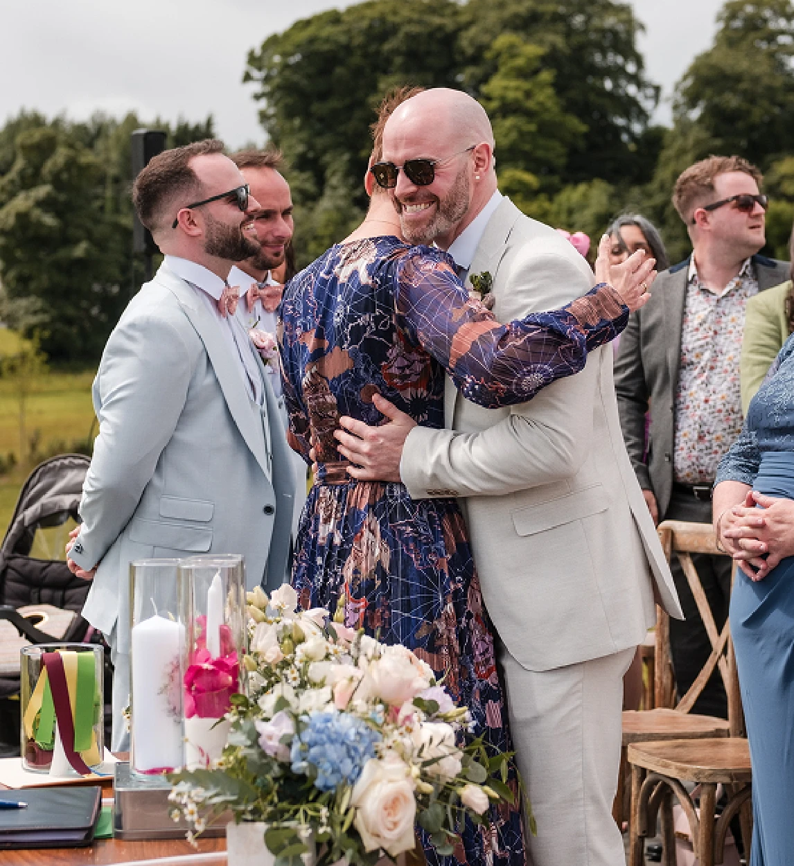 Rosemary Hartigan and the groom embracing beside the ceremony flowers, guests smiling behind them