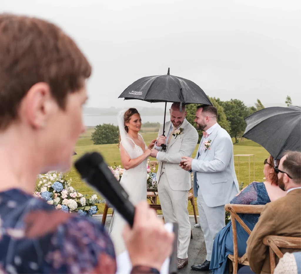 Rosemary Hartigan leading an outdoor ceremony in summer rain, the couple sheltering under an umbrella
